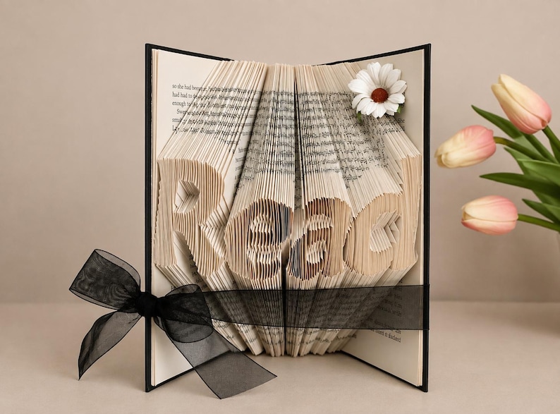 May include: A book with folded pages forming the word Read, featuring a black ribbon tied around the center and a white daisy flower accent on the top right corner. The book has a black cover and contains printed text on the interior pages.