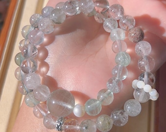 May include: Two beaded bracelets featuring clear and pale green stones. The bracelets are made of round beads of varying sizes, with a few larger beads and a small white pearl. The bracelets are held in a hand.