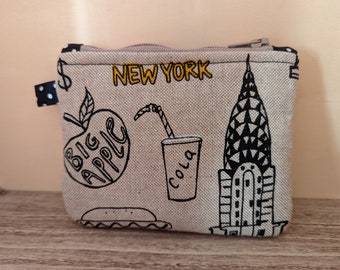 May include: A beige canvas zippered pouch with a black and white illustration of the Empire State Building, a red apple with the words "Big Apple", and a cup of cola. The pouch has the words "New York" printed in yellow.