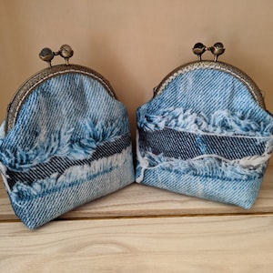 Könnte beinhalten: Zwei Münzbörsen aus hellblauem Denim mit Used-Details und ausgefransten Kanten. Jede Börse hat einen dekorativen Metallverschluss. Der Denim hat dunkelblaue Akzente.