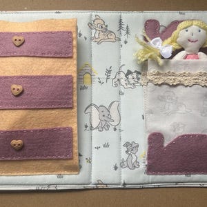 May include: A fabric quiet book page featuring a felt dresser with three mauve drawers and heart-shaped buttons on the left, and a small plush doll with yellow yarn hair tucked into a bed with a mauve headboard and footboard on the right.