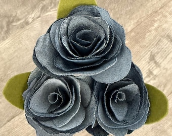 Pode incluir: Um arranjo floral artesanal com três rosas de ganga com construção detalhada das pétalas. As rosas são dispostas com folhas de feltro verde, criando uma peça decorativa única. As rosas de ganga são de cor azul média.