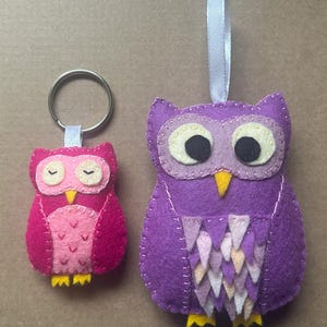 May include: Two felt owl ornaments. One is a keychain in shades of pink, with a yellow beak and feet. The other is a larger purple owl with a white ribbon for hanging, featuring a yellow beak and feet. Both have large eyes.
