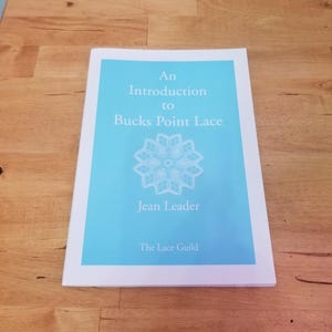 Puede incluir: Un libro titulado "An Introduction to Bucks Point Lace" con una cubierta azul claro y borde blanco. La portada presenta un diseño de encaje blanco y el nombre del autor, Jean Leader, junto con "The Lace Guild". El libro está sobre una superficie de madera.