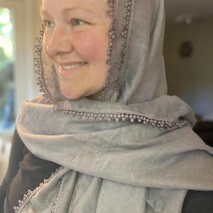 May include: A light gray scarf featuring a delicate pom-pom trim along the edges. The scarf is draped over the head and shoulders, showcasing a soft, woven texture and a consistent neutral color throughout the entire fabric and the decorative trim.