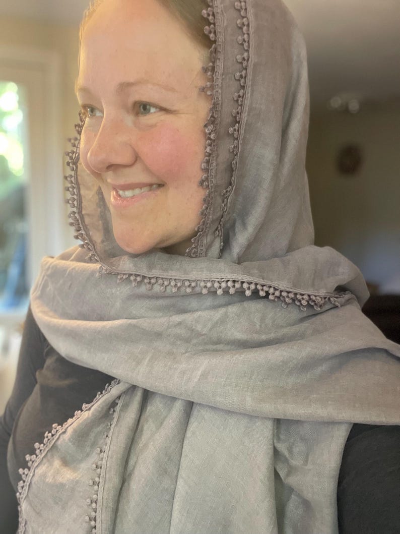 May include: A light gray scarf featuring a delicate pom-pom trim along the edges. The scarf is draped over the head and shoulders, showcasing a soft, woven texture and a consistent neutral color throughout the entire fabric and the decorative trim.