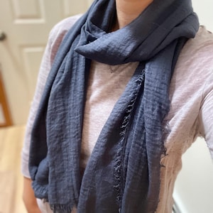 May include: A dark charcoal gray scarf with a crinkled, textured fabric finish and frayed edges. The scarf is draped around the neck, showcasing its lightweight, semi-sheer material and long rectangular shape with visible fringe detailing.