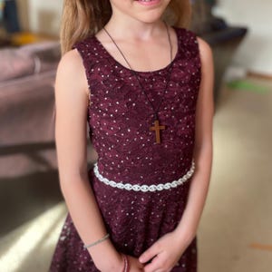 May include: A young girl wearing a burgundy lace dress with a white beaded belt and a wooden cross necklace.