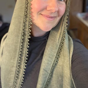 May include: A sage green head covering or scarf featuring a delicate, scalloped lace trim along the edges. The fabric has a subtle, textured appearance and is draped over the head, framing the face with its soft, muted green color and intricate border.