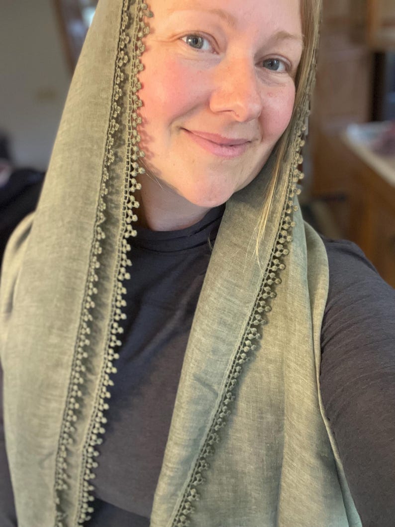 May include: A sage green head covering or scarf featuring a delicate, scalloped lace trim along the edges. The fabric has a subtle, textured appearance and is draped over the head, framing the face with its soft, muted green color and intricate border.