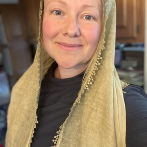 May include: A light olive green head covering made of a textured fabric with a delicate, scalloped lace trim along the edges. The fabric has a visible weave and drapes over the head and shoulders, worn over a solid black long-sleeved shirt.