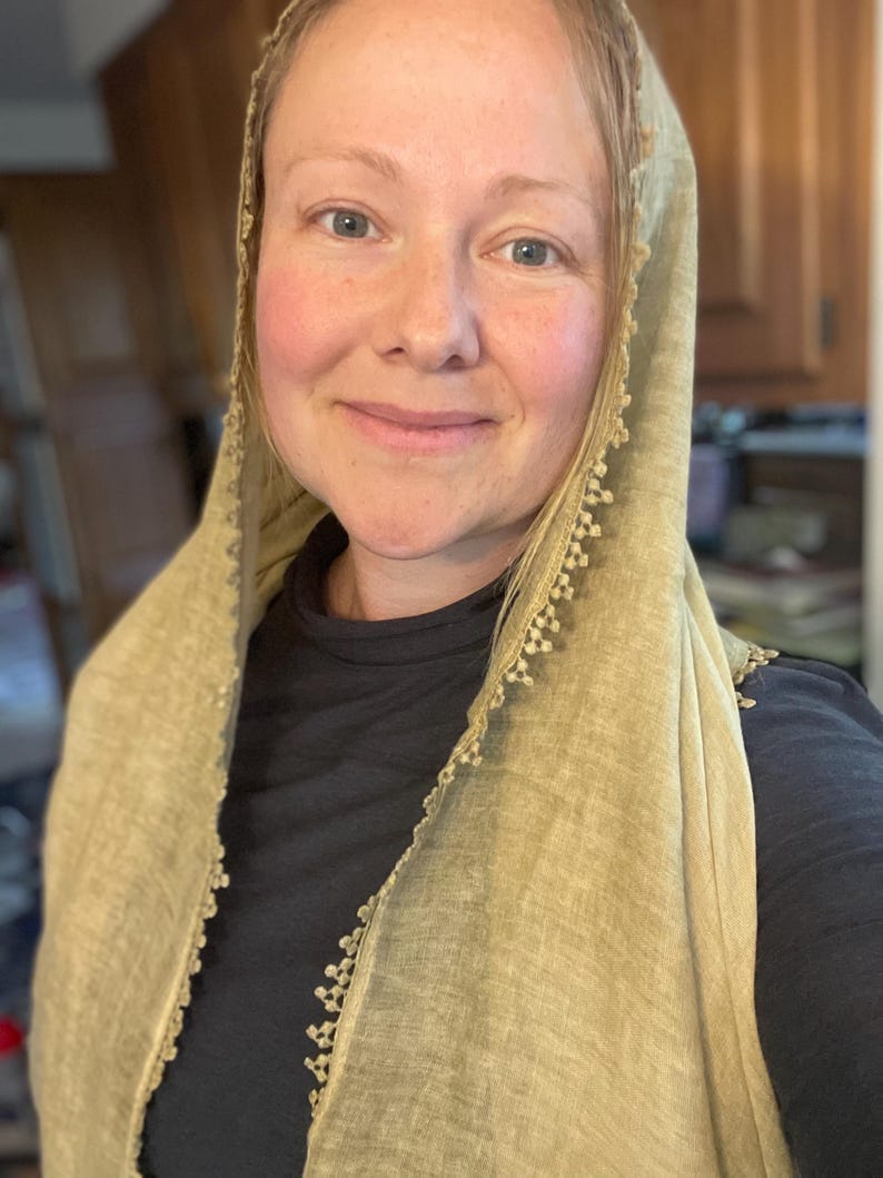 May include: A light olive green head covering made of a textured fabric with a delicate, scalloped lace trim along the edges. The fabric has a visible weave and drapes over the head and shoulders, worn over a solid black long-sleeved shirt.