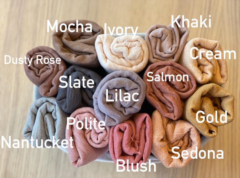 Op de afbeelding: Een verzameling opgerolde stoffen in verschillende kleuren, waaronder Mocha, Ivory, Khaki, Cream en Gold. Andere kleuren zijn Dusty Rose, Slate, Lilac, Salmon en Sedona. De stof is gerangschikt in een mand, met kleurnamen.