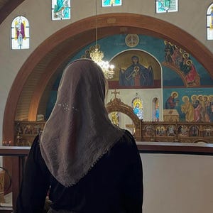 Op de afbeelding: Een persoon met een lichtbruine hoofdbedekking en een zwarte top staat in een kerk. Het interieur van de kerk heeft glas-in-loodramen, een muurschildering en een decoratieve kroonluchter. De persoon kijkt naar het altaar.