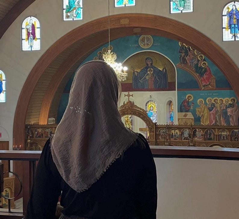 Op de afbeelding: Een persoon met een lichtbruine hoofdbedekking en een zwarte top staat in een kerk. Het interieur van de kerk heeft glas-in-loodramen, een muurschildering en een decoratieve kroonluchter. De persoon kijkt naar het altaar.