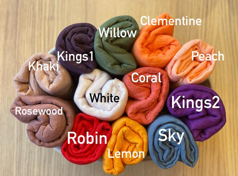 Op de afbeelding: Een verzameling opgerolde stoffen in verschillende kleuren, waaronder Khaki, Kings1, Willow, Clementine, Perzik, Kings2, Sky, Citroen, Robin, Rozenhout, Wit en Koraal. Elke stof is gelabeld met zijn kleurnaam.