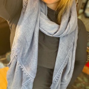 May include: A light blue scarf featuring a textured fabric and a delicate trim along the edges. The trim consists of small, evenly spaced beads or pom-poms attached to a decorative lace-like border that runs along the length of the scarf.