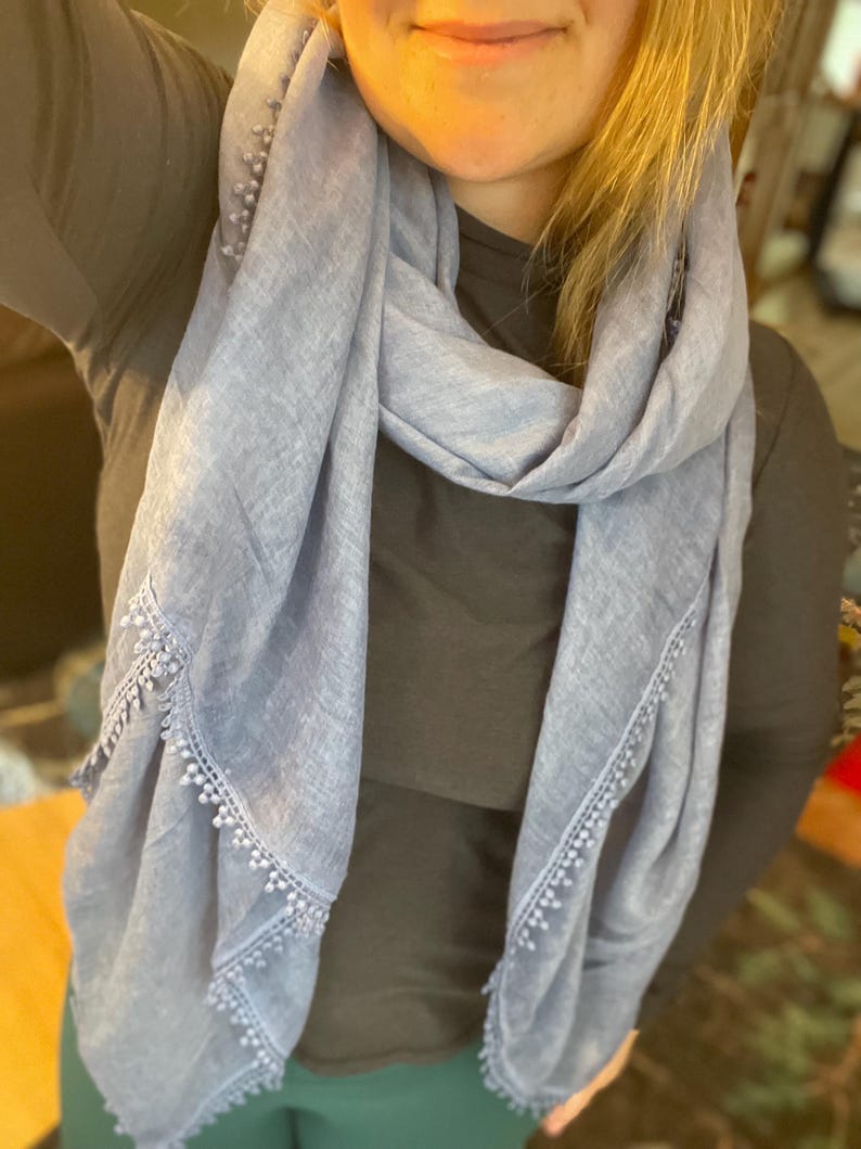 May include: A light blue scarf featuring a textured fabric and a delicate trim along the edges. The trim consists of small, evenly spaced beads or pom-poms attached to a decorative lace-like border that runs along the length of the scarf.