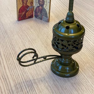May include: An olive green metal censer with a cross on top and a decorative handle. Two small religious icons are in the background, featuring images of religious figures. The censer has a decorative, openwork design.