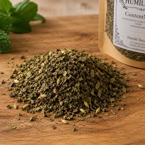 May include: A pile of loose leaf tea blend featuring dark green dried leaves and light tan seeds. Beside the pile is a brown paper pouch labeled Humili-Tea ContentMint, Handcrafted Loose Leaf Blend, Caffeine Free, Humili-Tea.com, Tewksbury, MA.