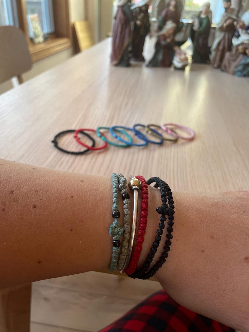 May include: A close-up of a person's wrist wearing several bracelets. The bracelets are made of different materials, including string, metal, and beads. The bracelets are in various colours, including black, red, blue, and green.