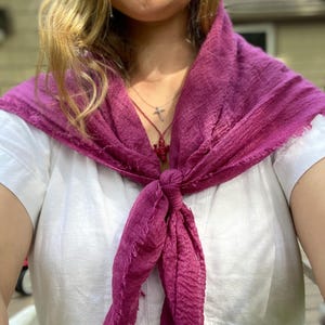 Op de afbeelding: Een magenta sjaal geknoopt om de nek, gedrapeerd over een witte jurk. De sjaal heeft een getextureerde uitstraling en gefranjerde randen. Een zilveren ketting met een kruisvormige hanger is zichtbaar.