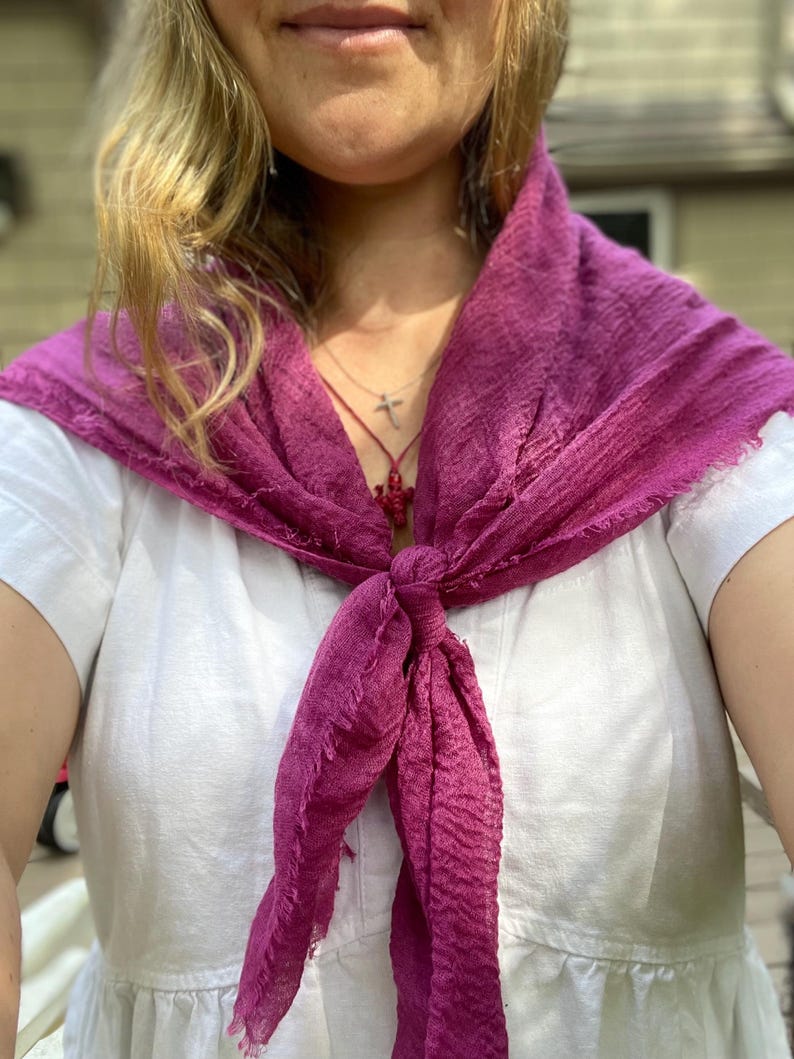 Op de afbeelding: Een magenta sjaal geknoopt om de nek, gedrapeerd over een witte jurk. De sjaal heeft een getextureerde uitstraling en gefranjerde randen. Een zilveren ketting met een kruisvormige hanger is zichtbaar.