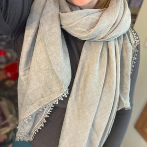 May include: A light gray scarf featuring a textured fabric finish and delicate white lace trim along the edges. The scarf is draped loosely around the neck, showcasing the soft material and the intricate, small-scale decorative border detail.