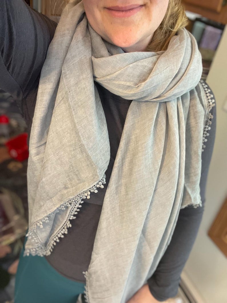 May include: A light gray scarf featuring a textured fabric finish and delicate white lace trim along the edges. The scarf is draped loosely around the neck, showcasing the soft material and the intricate, small-scale decorative border detail.