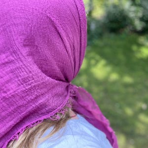 Op de afbeelding: Een levendige magenta hoofddoek, gedrapeerd en geknoopt, met een textuur. De stof is om het hoofd gewikkeld, met de uiteinden naar de zijkant. De achtergrond is een vage groene kleur, wat duidt op een buitenomgeving.