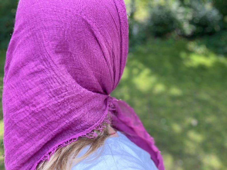 Op de afbeelding: Een levendige magenta hoofddoek, gedrapeerd en geknoopt, met een textuur. De stof is om het hoofd gewikkeld, met de uiteinden naar de zijkant. De achtergrond is een vage groene kleur, wat duidt op een buitenomgeving.