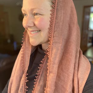 May include: A dusty rose-colored head covering made of a lightweight, semi-sheer fabric. The edges of the fabric are finished with a decorative, matching-colored trim featuring a small, repeating scalloped pattern along the border.