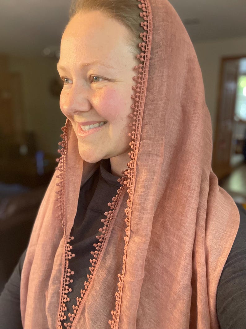May include: A dusty rose-colored head covering made of a lightweight, semi-sheer fabric. The edges of the fabric are finished with a decorative, matching-colored trim featuring a small, repeating scalloped pattern along the border.