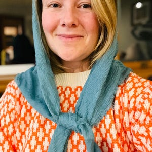 Op de afbeelding: Een vrouw draagt een lichtblauwe sjaal om de nek, gecombineerd met een oranje en witte trui met patroon. De trui heeft een geometrisch ontwerp. De vrouw glimlacht.