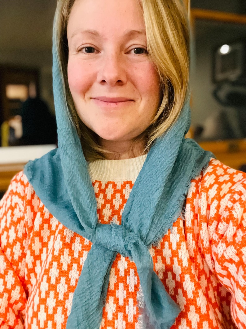 Op de afbeelding: Een vrouw draagt een lichtblauwe sjaal om de nek, gecombineerd met een oranje en witte trui met patroon. De trui heeft een geometrisch ontwerp. De vrouw glimlacht.