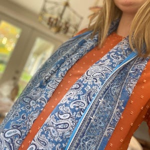 May include: A blue paisley scarf with white accents is draped over an orange top. The scarf features intricate patterns and a light blue border. The top has a textured, dotted design.
