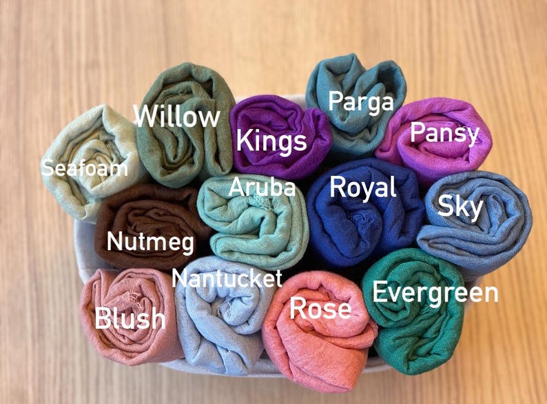 Op de afbeelding: Een verzameling opgerolde stoffen in verschillende kleuren, waaronder zeegroen, wilg, nootmuskaat, blush en roos. Elke rol is gelabeld met zijn kleurnaam, zoals "Kings", "Aruba" en "Evergreen". De stoffen zijn netjes gerangschikt in een witte container.