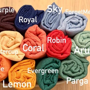 Op de afbeelding: Een verzameling opgerolde stoffen in verschillende kleuren, waaronder paars, blauw, oranje en groen. Elke rol is gelabeld met een kleurnaam, zoals "Royal", "Coral" en "Evergreen". De opstelling suggereert een display van sjaals of soortgelijke textielwaren.