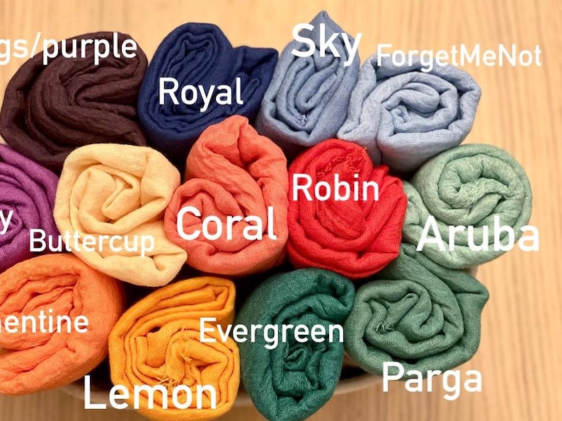 Op de afbeelding: Een verzameling opgerolde stoffen in verschillende kleuren, waaronder paars, blauw, oranje en groen. Elke rol is gelabeld met een kleurnaam, zoals "Royal", "Coral" en "Evergreen". De opstelling suggereert een display van sjaals of soortgelijke textielwaren.