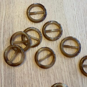 May include: A collection of brown, circular belt buckles with a rectangular bar across the center. The buckles are made of a translucent material and are scattered on a light-colored wooden surface.