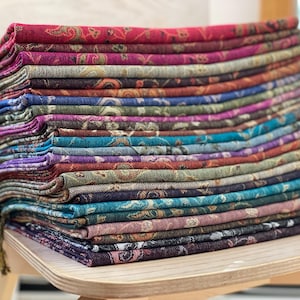 May include: A stack of colorful paisley-patterned scarves in shades of red, purple, blue, and green. The scarves are neatly folded and stacked on a wooden chair, showcasing a variety of colors and designs. The edges have a fringe.