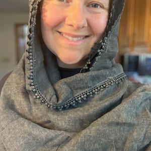 May include: A charcoal gray scarf with a textured fabric finish and a decorative trim featuring small, round pom-pom details along the edges. The scarf is draped loosely around the head and shoulders, showcasing the dark gray material and trim.
