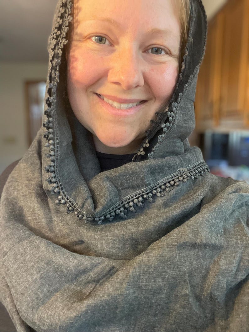 May include: A charcoal gray scarf with a textured fabric finish and a decorative trim featuring small, round pom-pom details along the edges. The scarf is draped loosely around the head and shoulders, showcasing the dark gray material and trim.