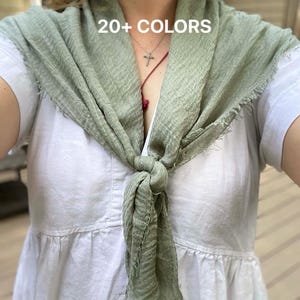 Op de afbeelding: Een saliegroene sjaal met een knoop en gerafelde randen. De sjaal wordt over een witte jurk gedragen. De tekst "20+ COLORS" is zichtbaar op de sjaal. Een veelzijdig accessoire.