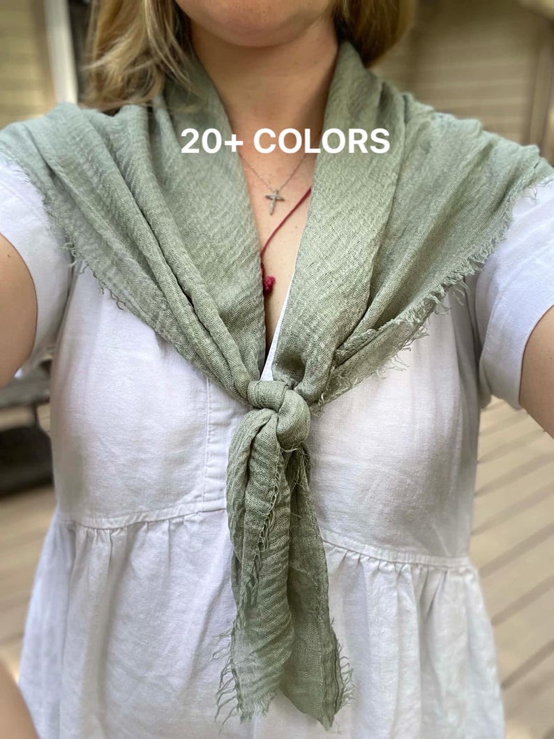 Op de afbeelding: Een saliegroene sjaal met een knoop en gerafelde randen. De sjaal wordt over een witte jurk gedragen. De tekst "20+ COLORS" is zichtbaar op de sjaal. Een veelzijdig accessoire.