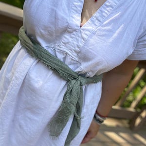 Op de afbeelding: Witte linnen jurk met een V-hals en korte mouwen, vastgebonden in de taille met een saliegroene sjaal. De sjaal is geknoopt. Een rode hanger hangt aan een ketting.