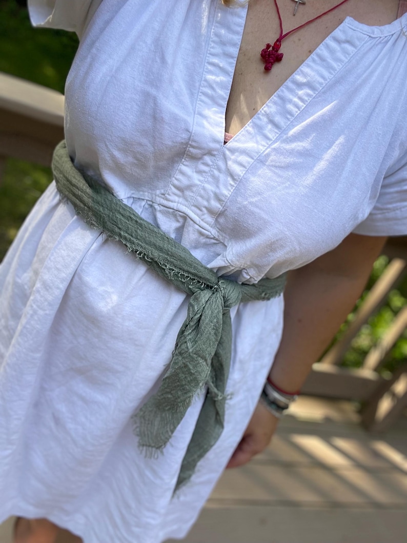 Op de afbeelding: Witte linnen jurk met een V-hals en korte mouwen, vastgebonden in de taille met een saliegroene sjaal. De sjaal is geknoopt. Een rode hanger hangt aan een ketting.