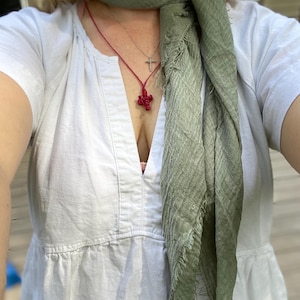 Op de afbeelding: Een vrouw draagt een witte V-hals jurk en een saliegroene sjaal. Een rode kruis hanger ketting en een zilveren kruis ketting zijn zichtbaar. De sjaal heeft een gefranjerde rand.