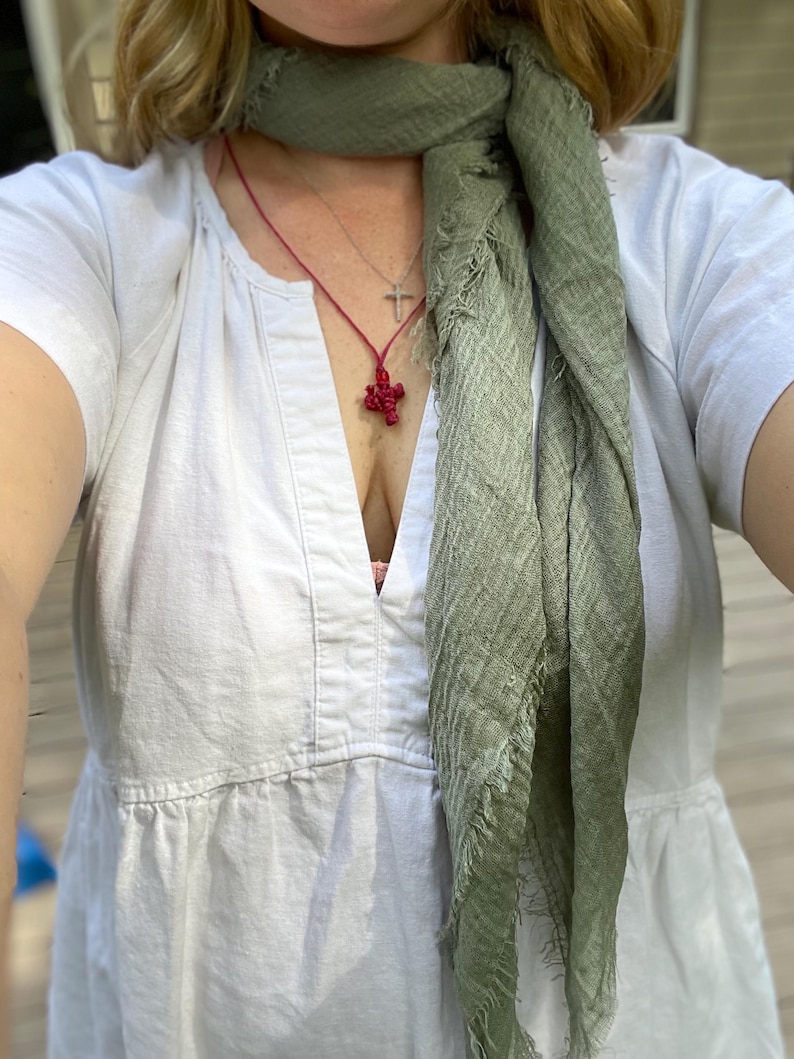 Op de afbeelding: Een vrouw draagt een witte V-hals jurk en een saliegroene sjaal. Een rode kruis hanger ketting en een zilveren kruis ketting zijn zichtbaar. De sjaal heeft een gefranjerde rand.