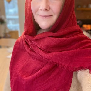 May include: A woman wearing a vibrant red scarf draped over her head and shoulders. The scarf has a textured appearance and a fringed edge. The background is softly blurred, suggesting an indoor setting.